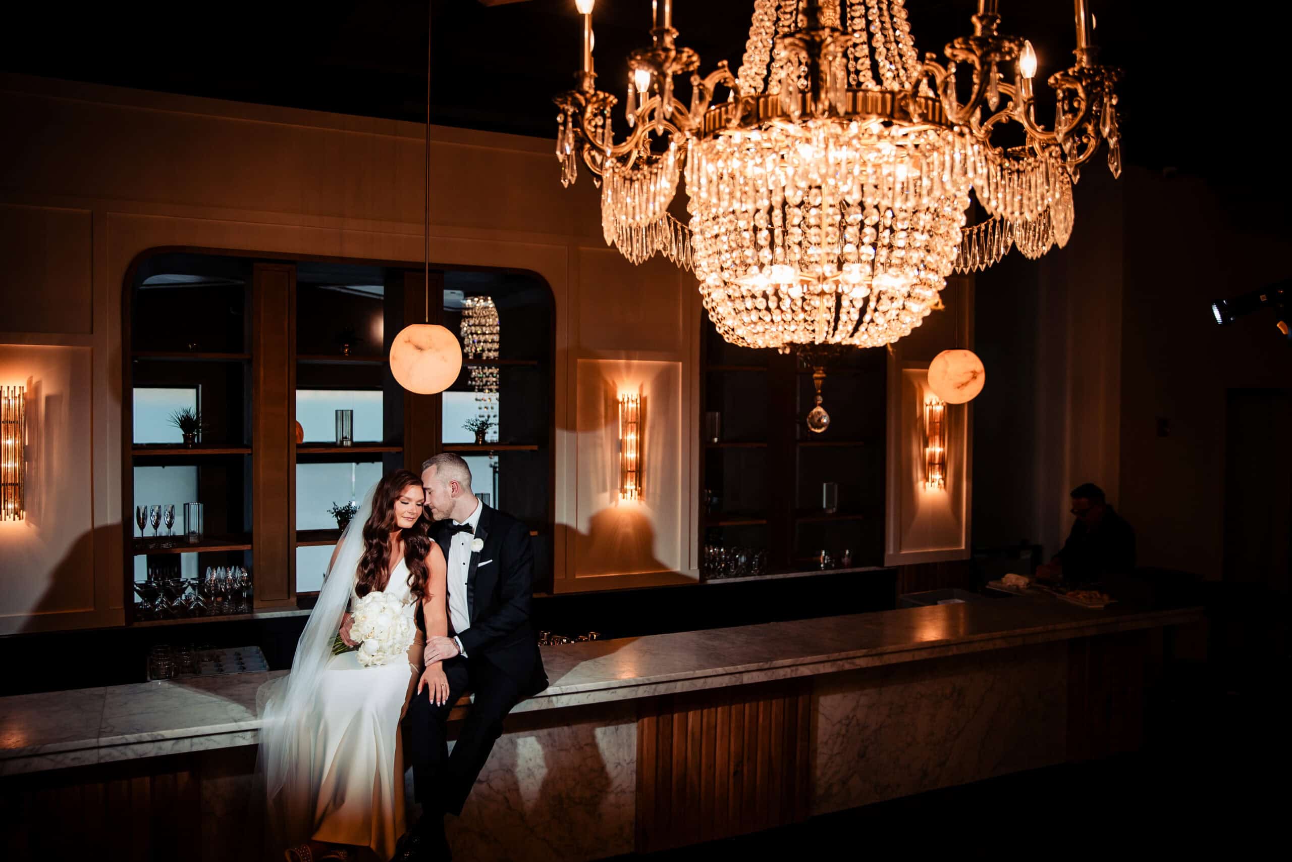 A bride and groom sit on a marble counter beneath a grand chandelier at Tendenza. The bride, in a white dress and veil, holds a bouquet, while the groom, in a black suit, kisses her forehead. They are in a dimly lit, elegant room with shelves and warm lighting—a perfect moment captured by Cescaphe Wedding Photography.